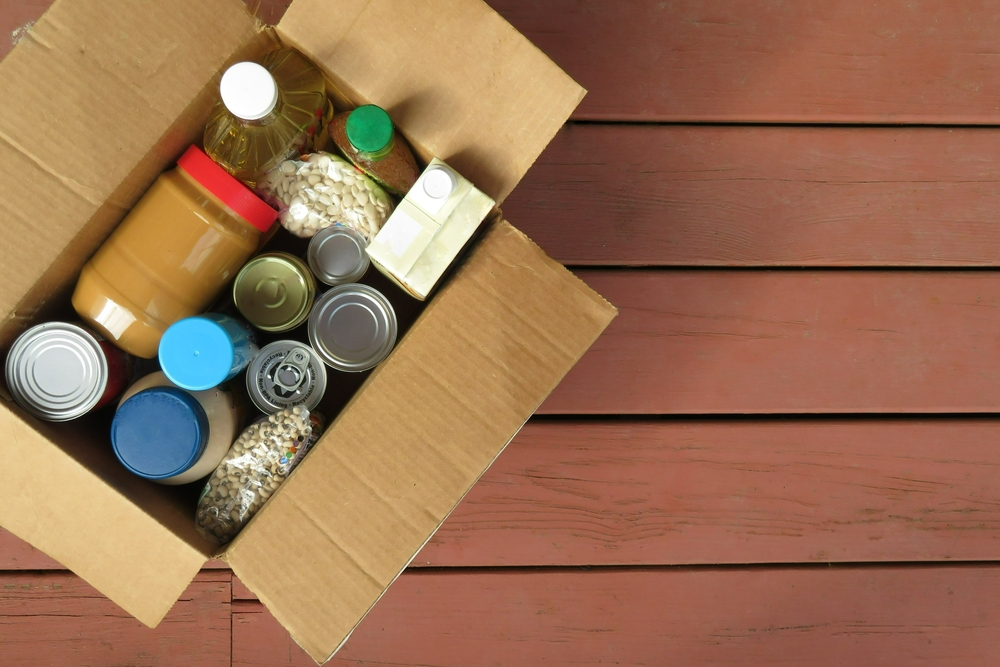 A box of donated canned goods and non-perishable foods for a food pantry for the poor sitting on red, wood panel background.