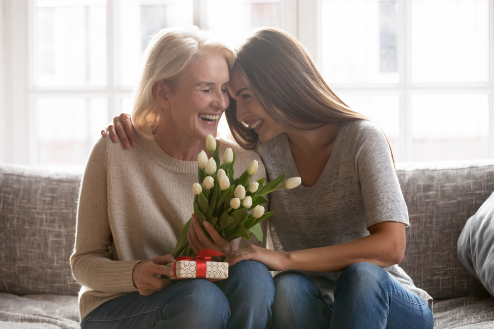 Loving young adult female child congratulate excited elderly mother with birthday anniversary at home. Smiling caring grownup millennial daughter present gift flowers to old mom on women s day.